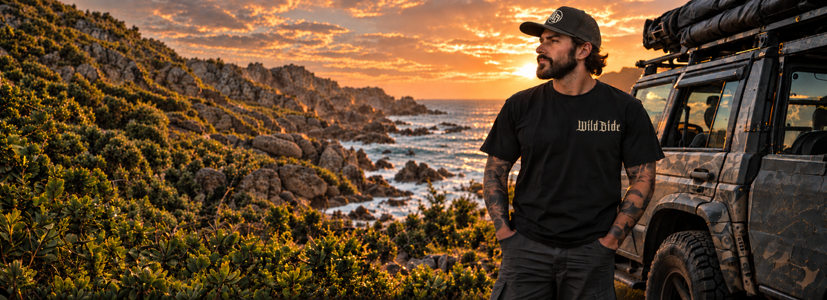 Man wearing Wild Side HQ apparel beside a 4WD on a coastal track at sunset